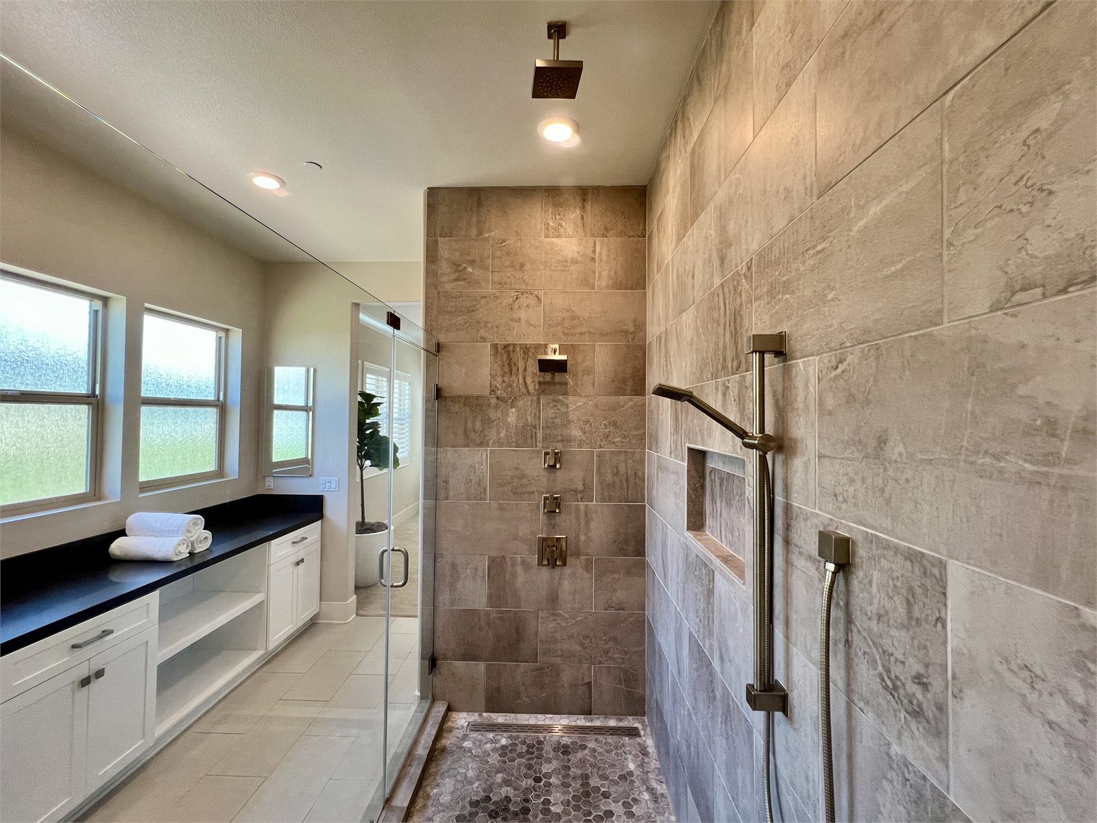 Master bathroom with a large walk-in shower in warm tan stone tile and a long white vanity with a black countertop