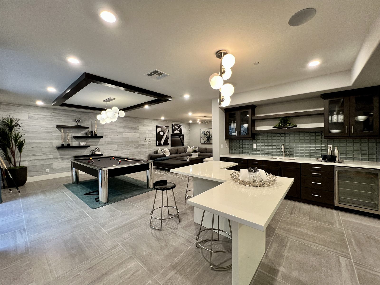 Lower-level lounge with a pool table, sectional seating, and a wet bar finished in white marble-look tile